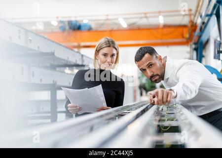 Businessman and woman with papers examining metal rods in factory hall Stock Photo