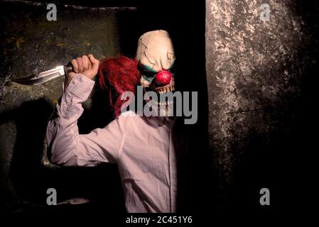 scary clown, in the shadows, looking eerily at the camera from behind an abstract wall. Halloween concept Stock Photo