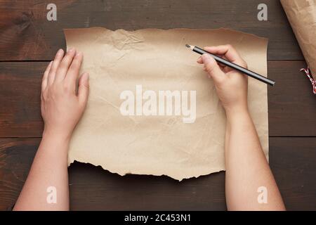 empty torn sheet of brown craft paper and two hands with a black wooden pencil, wooden table, top view, place for an inscription Stock Photo