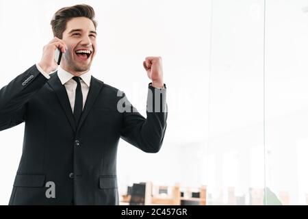Image of excited young businessman wearing black suit talking cellphone and making winner gesture in office Stock Photo