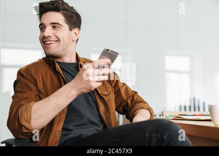 Image of handsome joyful young man smiling and using cellphone while drinking coffee at office Stock Photo