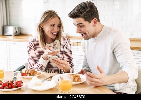 Portrait of young happy couple using smartphone while having breakfast in cozy kitchen at home Stock Photo