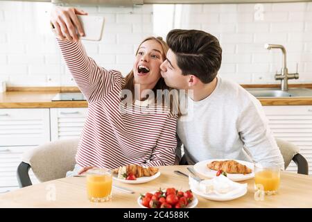Portrait of young excited couple taking selfie on smartphone while having breakfast in cozy kitchen at home Stock Photo