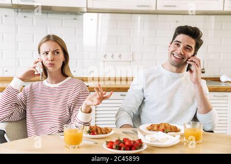 Portrait of young caucasian couple talking on smartphones while having breakfast in cozy kitchen at home Stock Photo