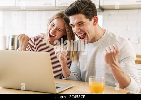 Portrait of young excited couple using laptop and making winner gesture while having breakfast in cozy kitchen at home Stock Photo