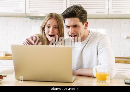 Portrait of young surprised couple using laptop while having breakfast in cozy kitchen at home Stock Photo