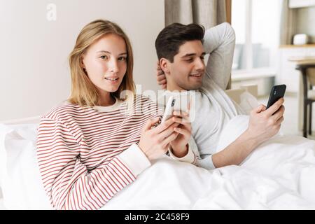 Portrait of beautiful happy couple smiling and using smartphones while lying in bed at home Stock Photo