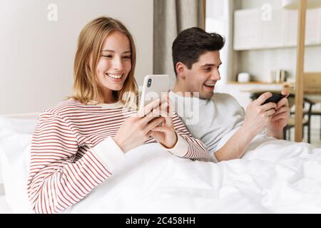 Portrait of beautiful happy couple smiling and using smartphones while lying in bed at home Stock Photo