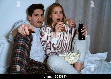 Portrait of shocked caucasian couple watching tv and eating popcorn while sitting in bed at home Stock Photo