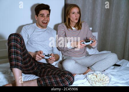 Portrait of excited cute couple holding joysticks while playing video games in bed at home Stock Photo
