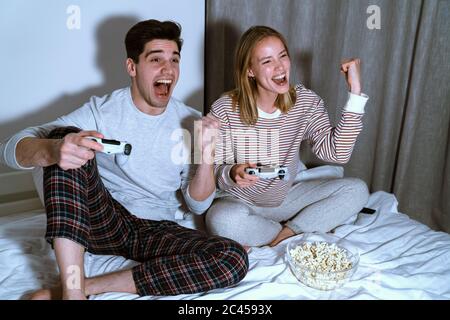 Portrait of delighted cute couple holding joysticks and making winner gesture while playing video games in bed Stock Photo