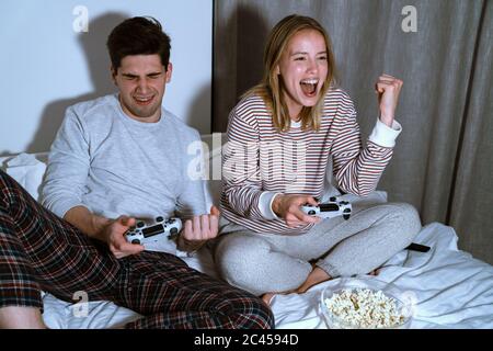 Portrait of emotional cute couple holding joysticks and making winner gesture while playing video games in bed Stock Photo