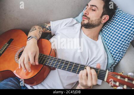 Young bearded man lying on sofa, playing acoustic guitar. Stock Photo