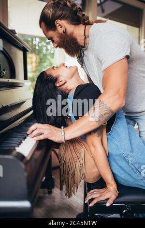 Bearded tattooed man with long brunette hair leaning over smiling woman, playing piano. Stock Photo