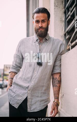 Portrait of bearded young man with brown hair, with tattoos on arms, wearing grey shirt, leaning against wall. Stock Photo