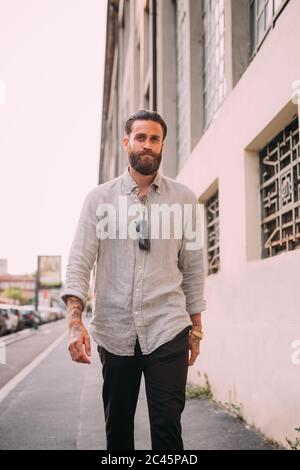 Portrait of bearded young man with brown hair, with tattoos on arms, wearing grey shirt, walking down street. Stock Photo