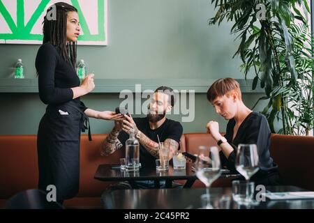 Young woman with short hair and bearded young man sitting in a bar, using mobile phone to pay. Stock Photo