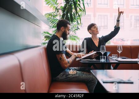 Young couple sitting at a table in a bar, woman beckoning for the bill. Stock Photo