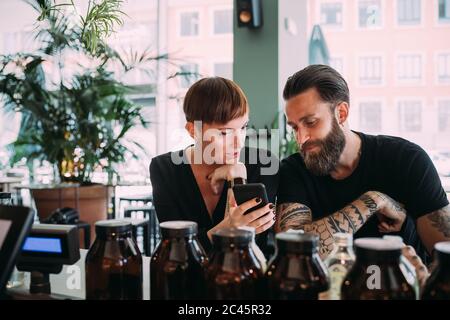 Bearded young man with brown hair and tattoos and young woman with short hair sitting in a bar, looking at mobile phone. Stock Photo