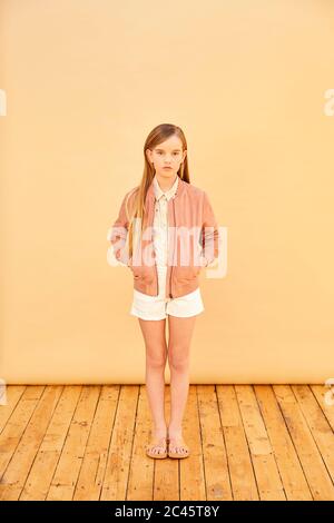 Portrait of girl with long blond hair wearing shorts, shirt and pink jacket, on pale yellow background. Stock Photo