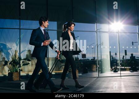 Businessman and businesswoman with wheeled luggage passing glass building, Malpensa, Milan Stock Photo
