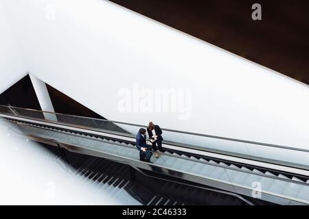 Businessman and businesswoman with wheeled luggage on escalator in hotel building Stock Photo