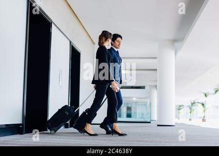 Businessman and businesswoman with wheeled luggage exiting hotel escalators Stock Photo