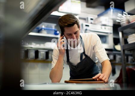 Young male chef reading digital tablet and talking on smartphone in kitchen Stock Photo
