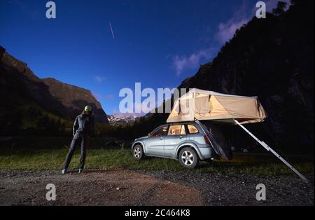 Grossglockner, Austria - July 14, 2019: Back view of hiker standing on grass near car with camping tent on rooftop and admiring the view of rocky hills at night Stock Photo