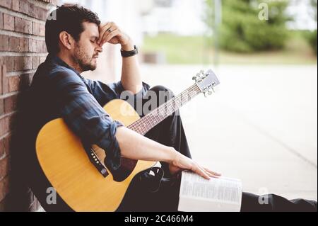 Sad man sitting on the ground holding a book and a guitar Stock Photo