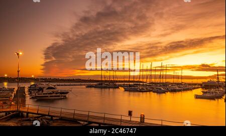 A view of the marina of Santa Maria di Leuca at red and orange sunset. Sailboats and yachts moored on the docks of the port. Wooden walkways lead to h Stock Photo