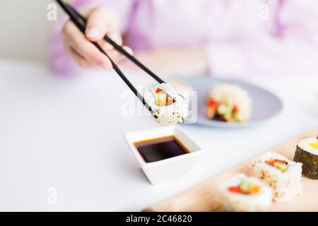 Close up of woman holding sushi roll with chopsticks above bowl full of soy sauce. Healthy traditional japanese meal concept. Stock Photo