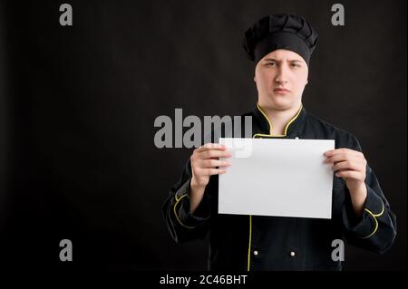 Portrait of young male dressed in a black chef suit holding a white paper, upset posing on a black isolated background with copy space advertising are Stock Photo