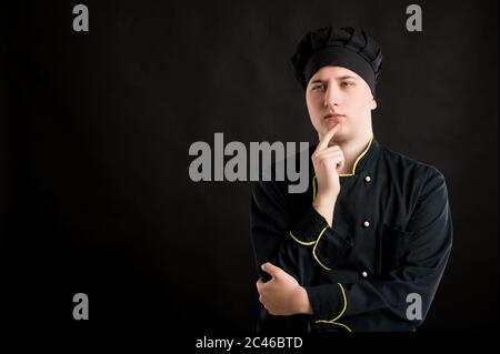 Portrait of young male dressed in a black chef suit holding hand under her chin, having doubtful posing on a black isolated background with copy space Stock Photo