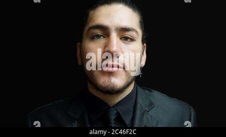 Close-up portrait of young latino man with black suit, black shirt, black tie seen from front on black background Stock Photo
