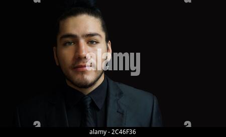 Portrait of a young latin man wearing earring with black suit, black shirt and black tie on black background Stock Photo