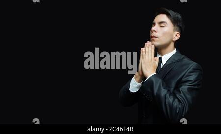 Young latin man in black suit, white shirt, black tie with his hands in prayer position on black background Stock Photo