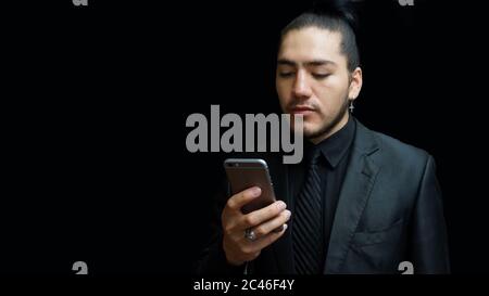 Young latin man in black suit, black shirt, black tie looking at a message on his cell phone on black background Stock Photo