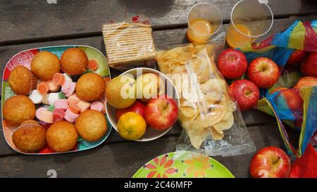 Red apples, pears, tangerines, salt crackers, plastic bag with chips and a plate with cupcakes and candies on a rustic wooden table Stock Photo