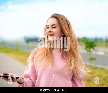 A young positive girl with a happy smile listens to music with headphones on her phone while walking down the street in the summer. Stock Photo