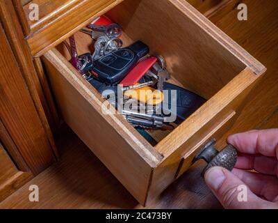 Close overhead shot of man’s fingers on the knob of a small open drawer in an old pine unit, holding an assortment of vehicle  door and ignition keys. Stock Photo