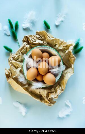 An Easter still life flat lay photo of a bowl of brown eggs on crumpled craft paper, white feathers and dry green plants as a decor. Light blue backgr Stock Photo