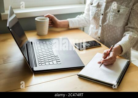 Woman's hands holding a pen over a notebook and a cup. Using laptop on desk in home interior. Remote work. Freelance. Stock Photo