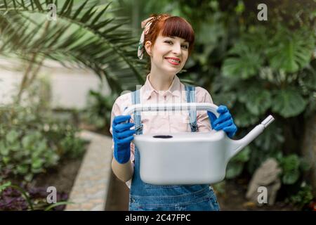 Gardening and greenhouse concept. Young cheerful red haired woman in jeans overalls, posing with gray water can for watering plants in floral Stock Photo