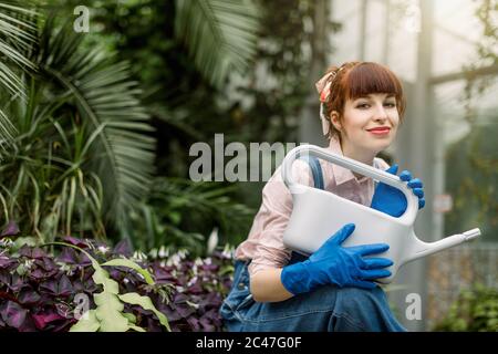 A beautiful woman in jeans overalls, pink shirt and blue gloves, in her garden or greenhouse, posing to camera with gray watering can Concept of Stock Photo