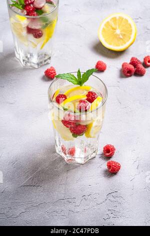 Fresh cold ice water with lemon, raspberry fruits and mint leaf in two faceted glass on stone concrete background, summer diet beverage, angle view Stock Photo