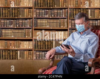 Senior caucasian man with a face mask against coronavirus reading an ebook tablet in old fashioned library of antique books Stock Photo