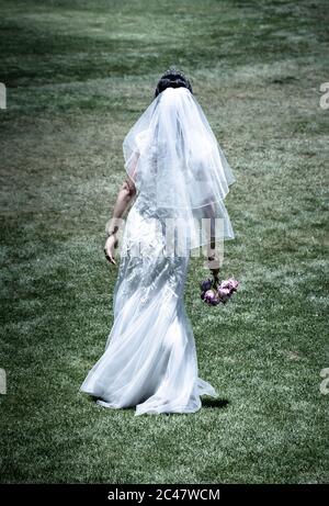 Isolated Rear view of bride in long wedding gown with veil holding a bouquet while walking outside in garden Stock Photo