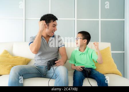 Asian family father and his son to playing video games with joysticks and arm raised to winning in game while sitting in sofa in living room at home, Stock Photo
