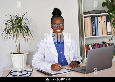 Smiling african female doctor wear glasses looking at camera sit at workplace. Stock Photo